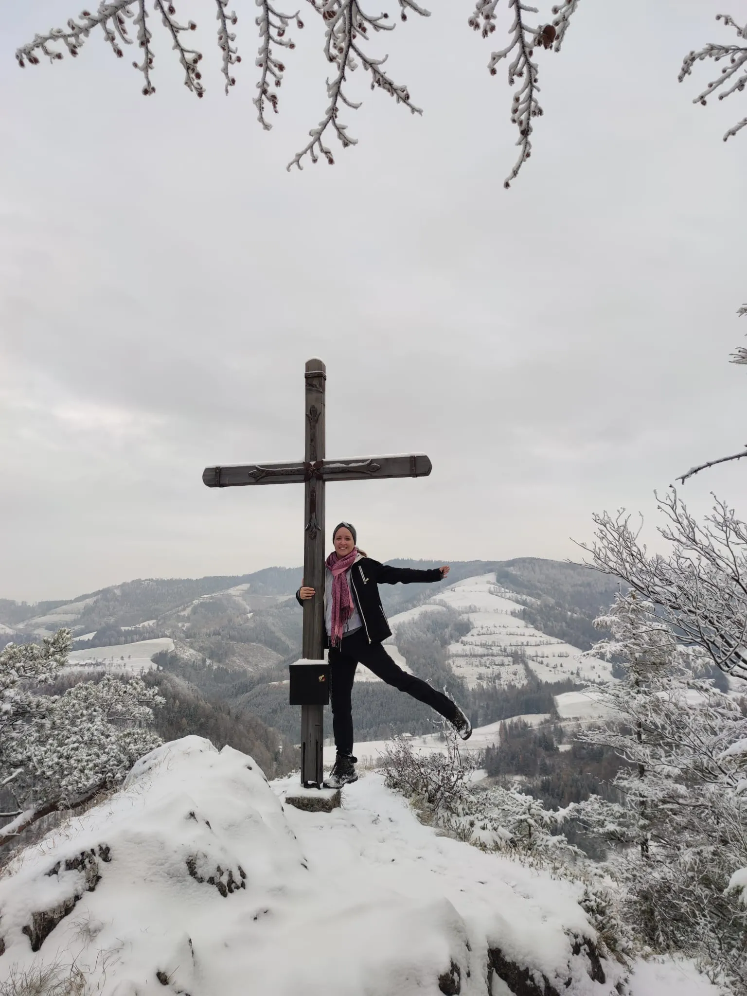 Jennifer ausgelassen am verschneiten Gipfelkreuz, dahinter die winterlichen Voralpenhügel